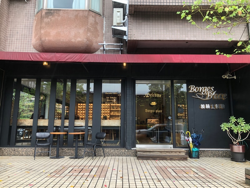 波赫士領地咖啡館明水店，玻璃門面與金色字體交織出靜謐氛圍，綠意點綴街角，彷彿一場文學與感官的邀請。